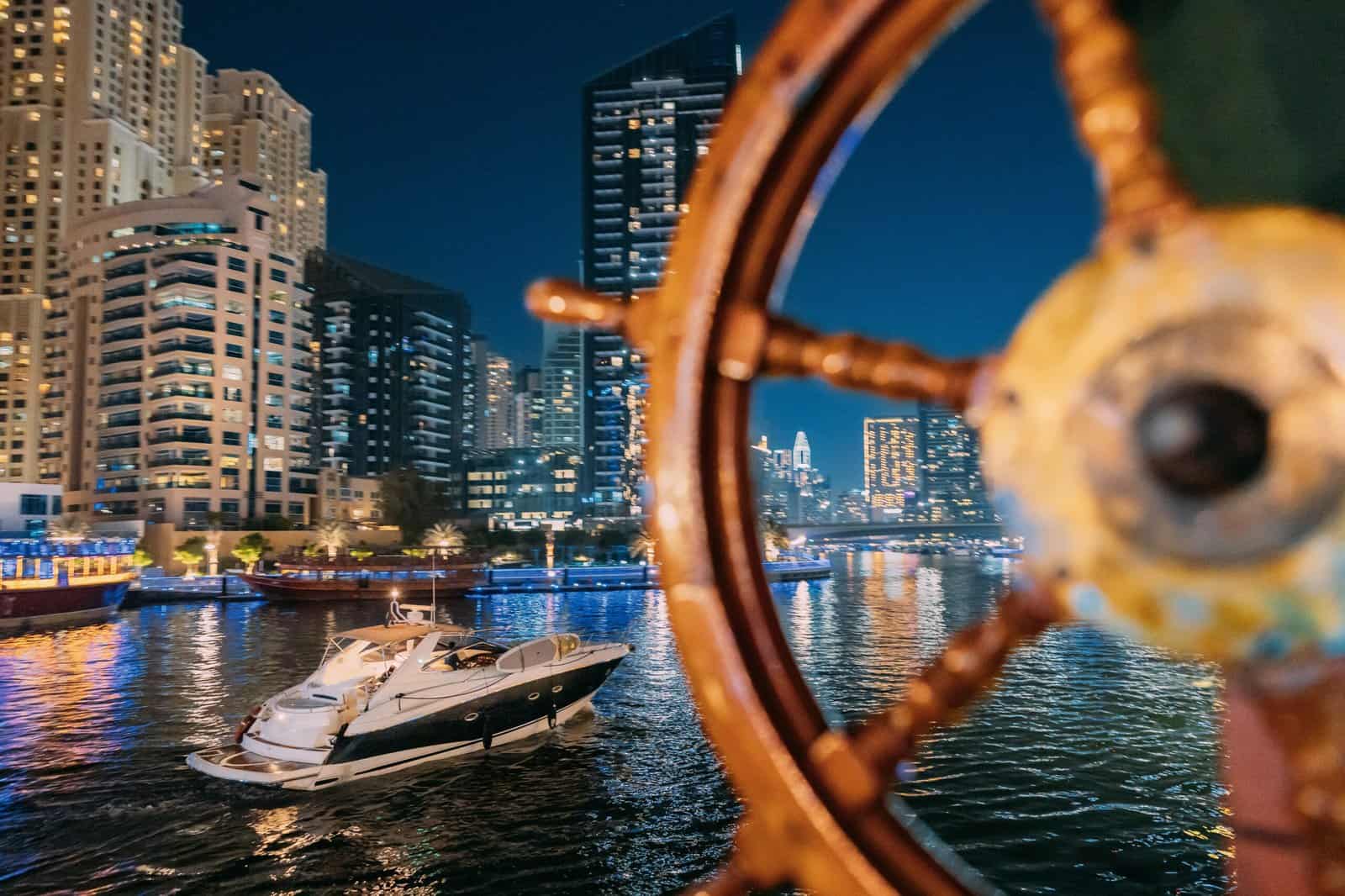 Steering Wheel On Ship At Skyline Background. Night View Of City Skyline From Tourist Boat, Sightseeing Boat Sailing On Dubai Marina. Night Walk Of Dubai Marina Is District in Dubai, United Arab Emirates. Nighttime
